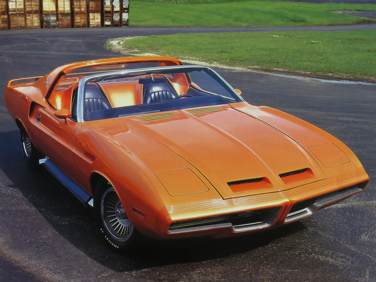 A bright orange classic convertible car with a sleek, aerodynamic design is parked on an asphalt surface near grass, featuring pop-up headlights and a sporty, retro appearance.