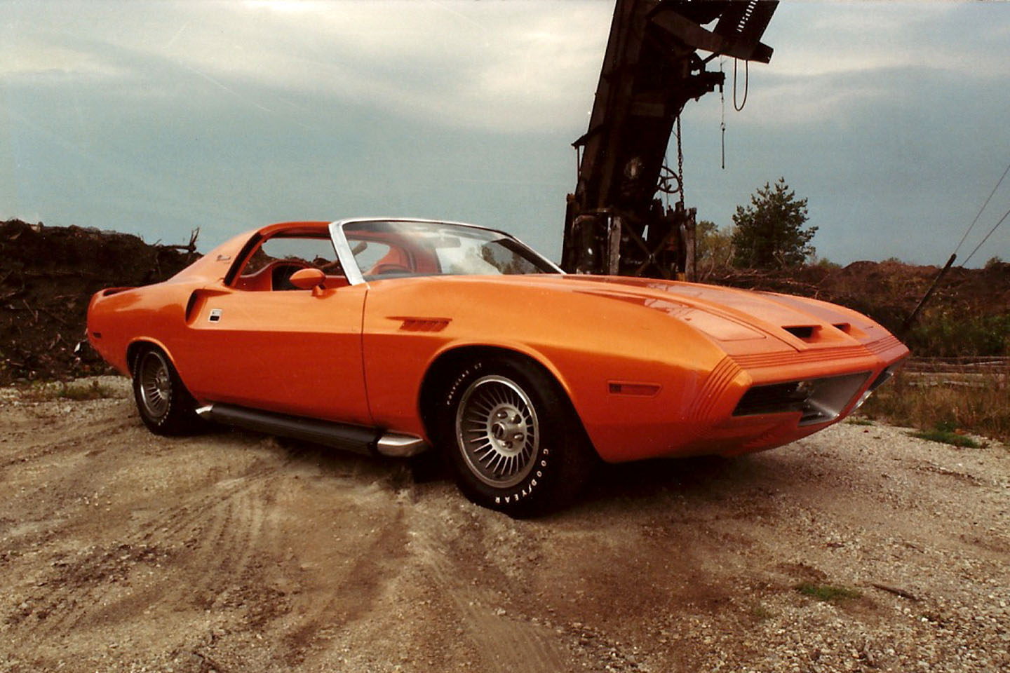 An orange classic muscle car with a t-top roof is parked on a dirt surface near industrial machinery, under a cloudy sky.