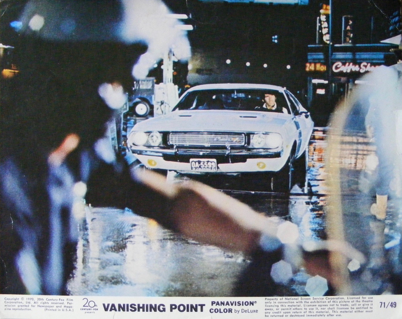 A white muscle car is stopped at night in a wet, neon-lit street while a police officer stands in the foreground, partially out of focus. The scene is from the film Vanishing Point.