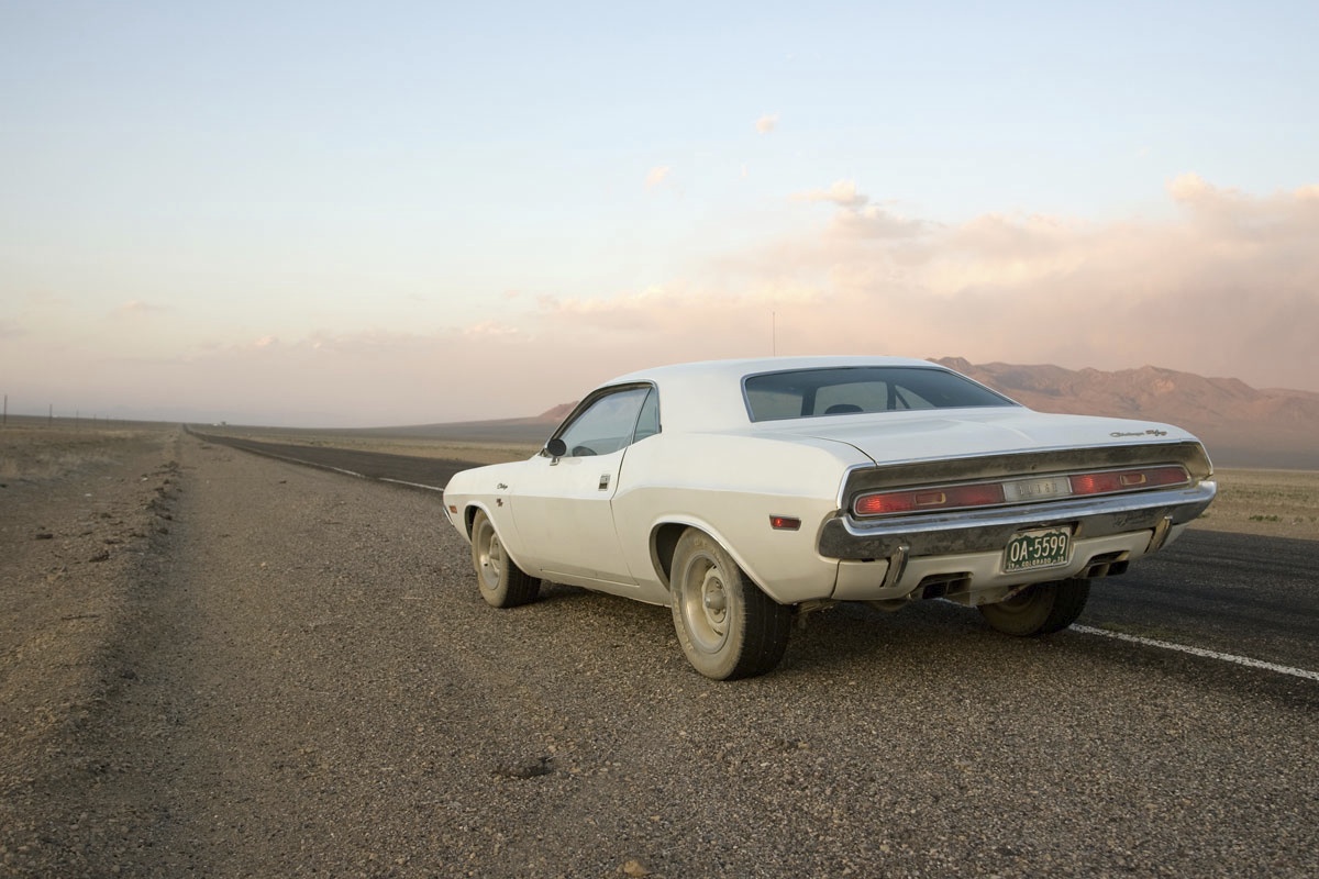 A classic white muscle car is parked on the side of a deserted road in a vast, open landscape with mountains in the distance under a clear sky.