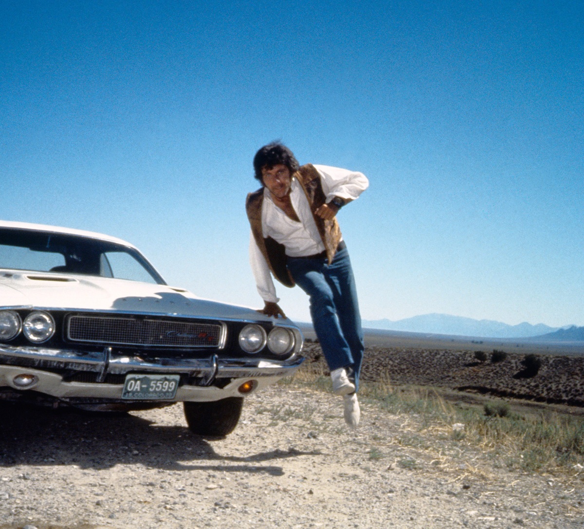 A man in jeans, a white shirt, and a brown vest jumps off the hood of a classic white car parked on a dirt roadside, with mountains and a clear blue sky in the background.