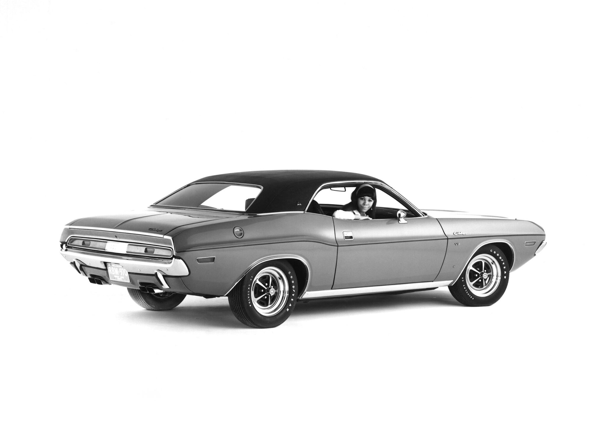 A black and white photo of a vintage muscle car, viewed from the rear side, with a woman sitting in the driver’s seat and smiling out the open window. The car is parked against a plain white background.