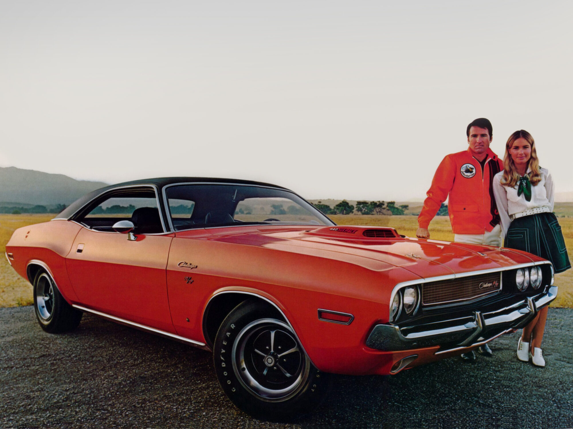 A man and a woman stand beside an orange classic Dodge Challenger parked on a road, with grass and mountains in the background under a clear sky.
