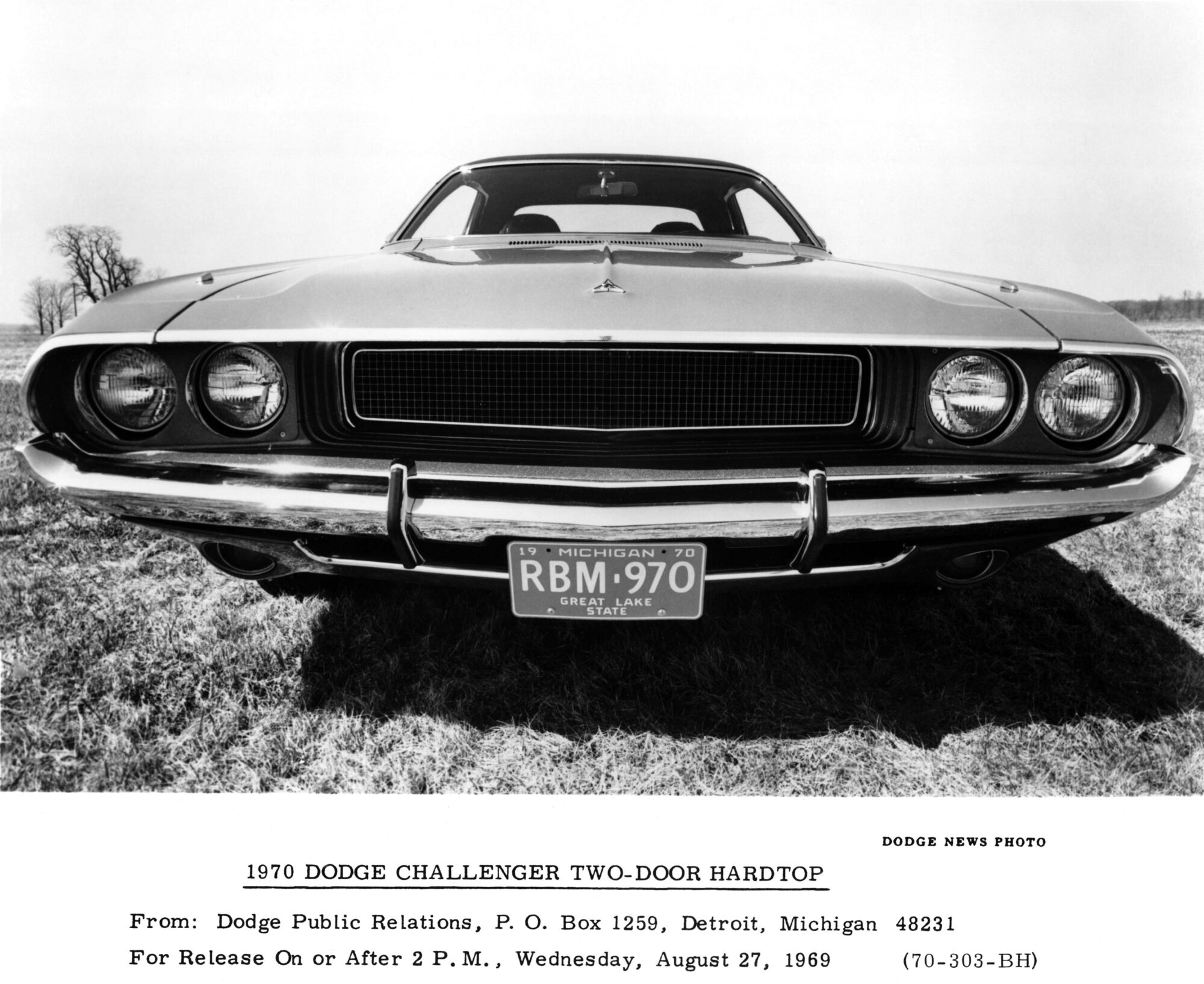 Black and white photo of a 1970 Dodge Challenger two-door hardtop, viewed from the front with a low, dramatic angle; license plate reads RBM 970. Grass and two trees are visible in the background.