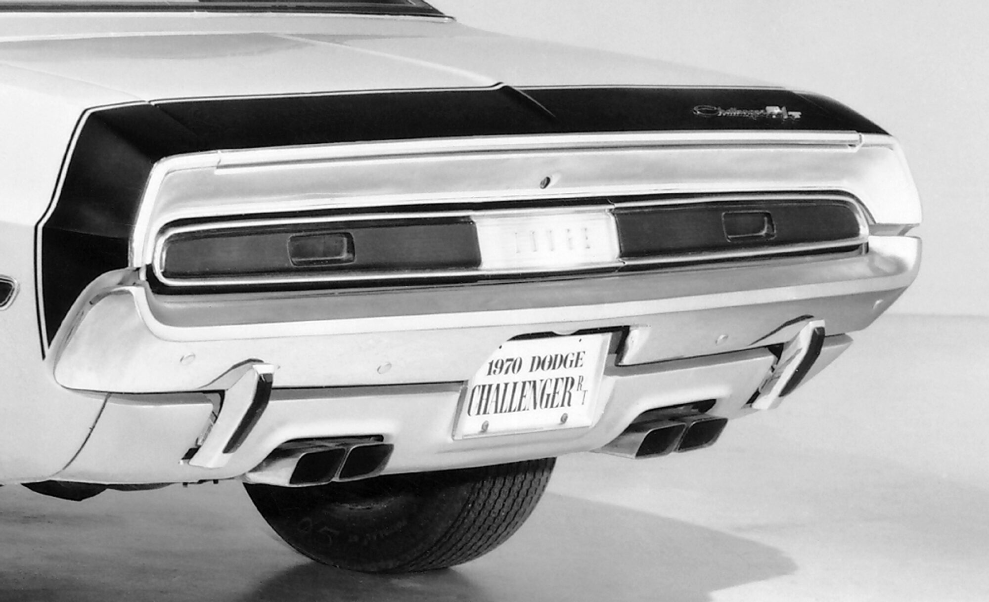 Black and white photo showing the rear view of a vintage car with dual exhausts, rectangular taillights, and a license plate that reads “1970 Dodge Challenger R/T.”.