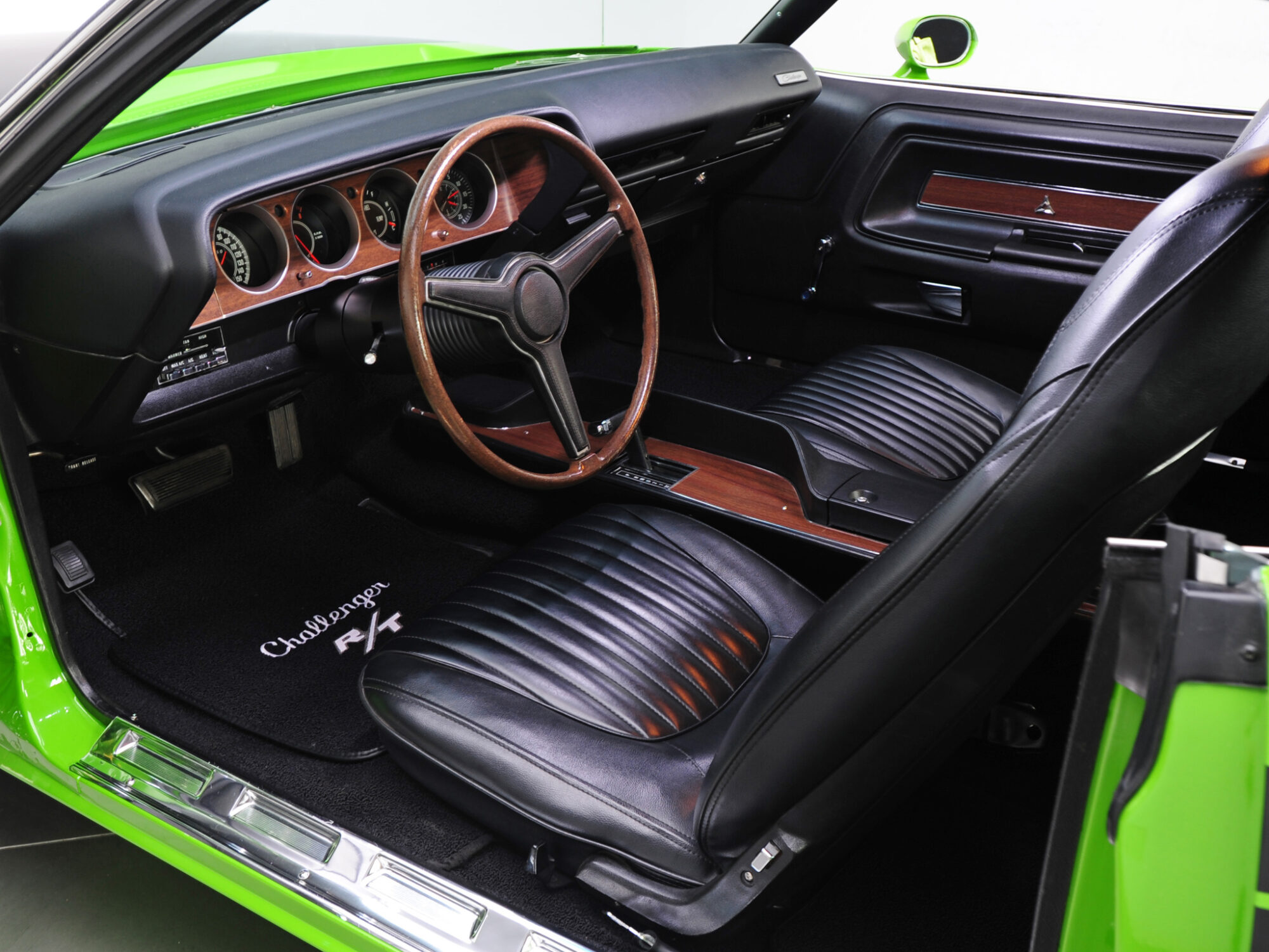 Interior of a classic car with black leather seats, a wood-trim steering wheel, wood-panel dashboard, and Challenger R/T floor mats. The exterior is bright green, visible on the door frame.