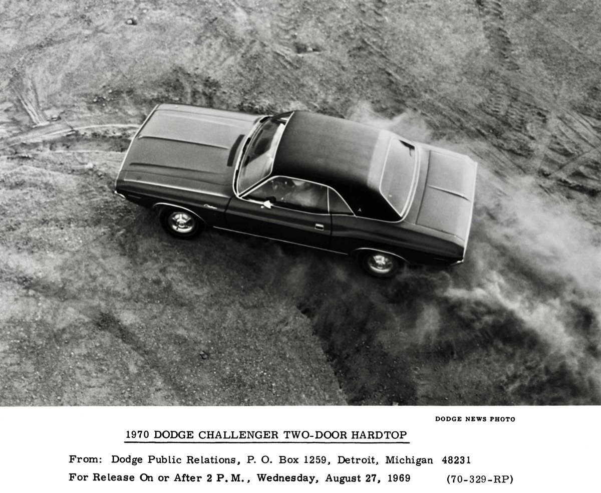A black-and-white overhead photo of a 1970 Dodge Challenger two-door hardtop driving on a dirt surface, kicking up dust, with a press release caption below the image.