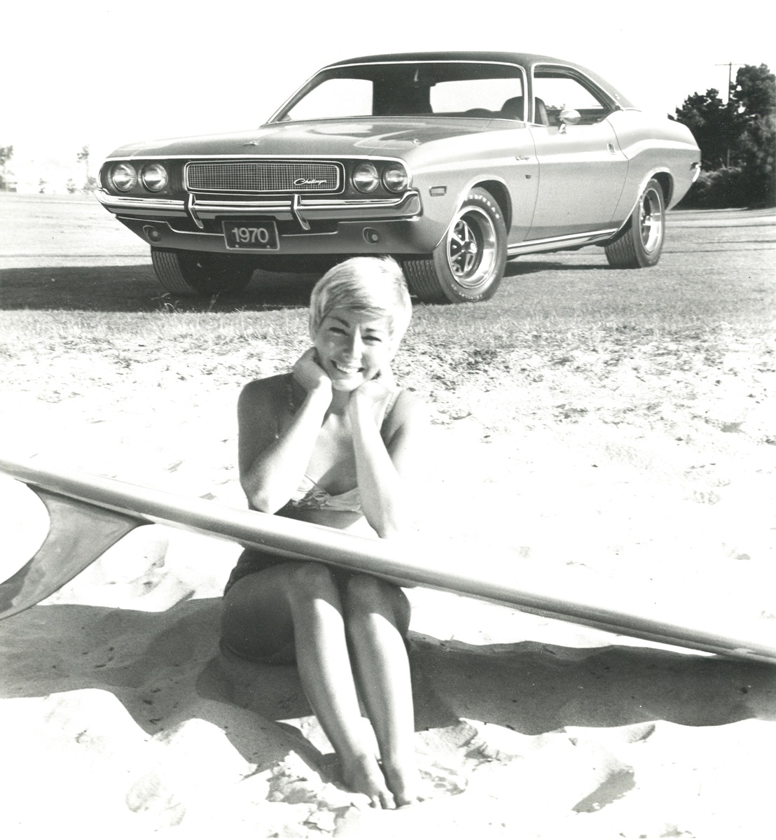 A woman in a swimsuit sits on sandy ground beside a surfboard, smiling at the camera, with a classic 1970 Dodge Challenger car parked behind her.