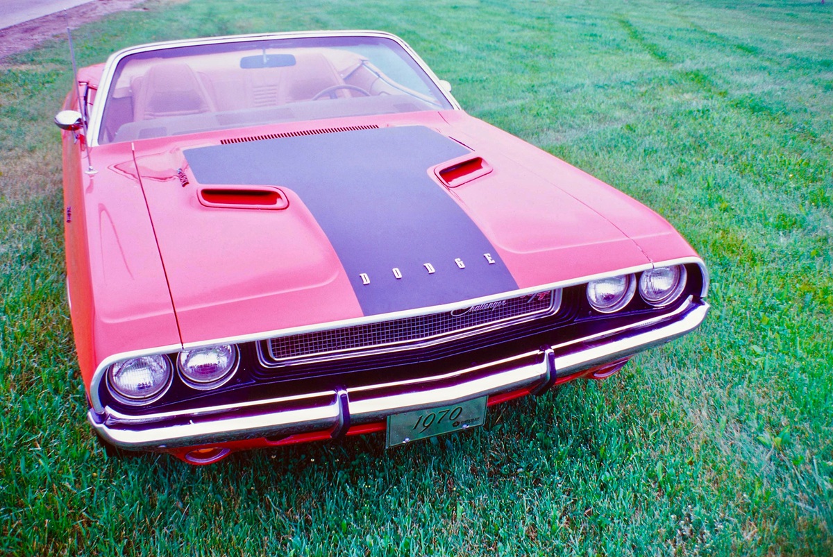 A red 1970 Dodge Challenger convertible with a black racing stripe on the hood is parked on green grass. The cars front grille, round headlights, and classic design are clearly visible.