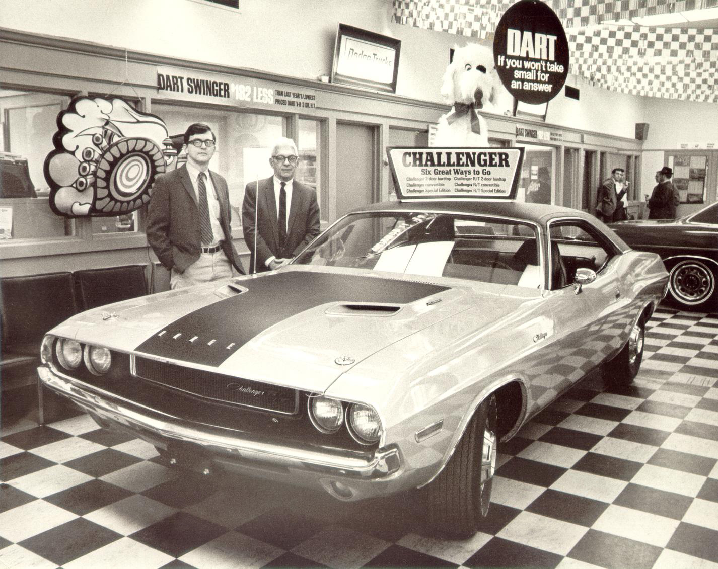 Two men in suits stand next to a vintage Dodge Challenger in a showroom with checkered floors. Signs for Dart Swinger and Challenger are visible, along with a large Snoopy figure above the car.