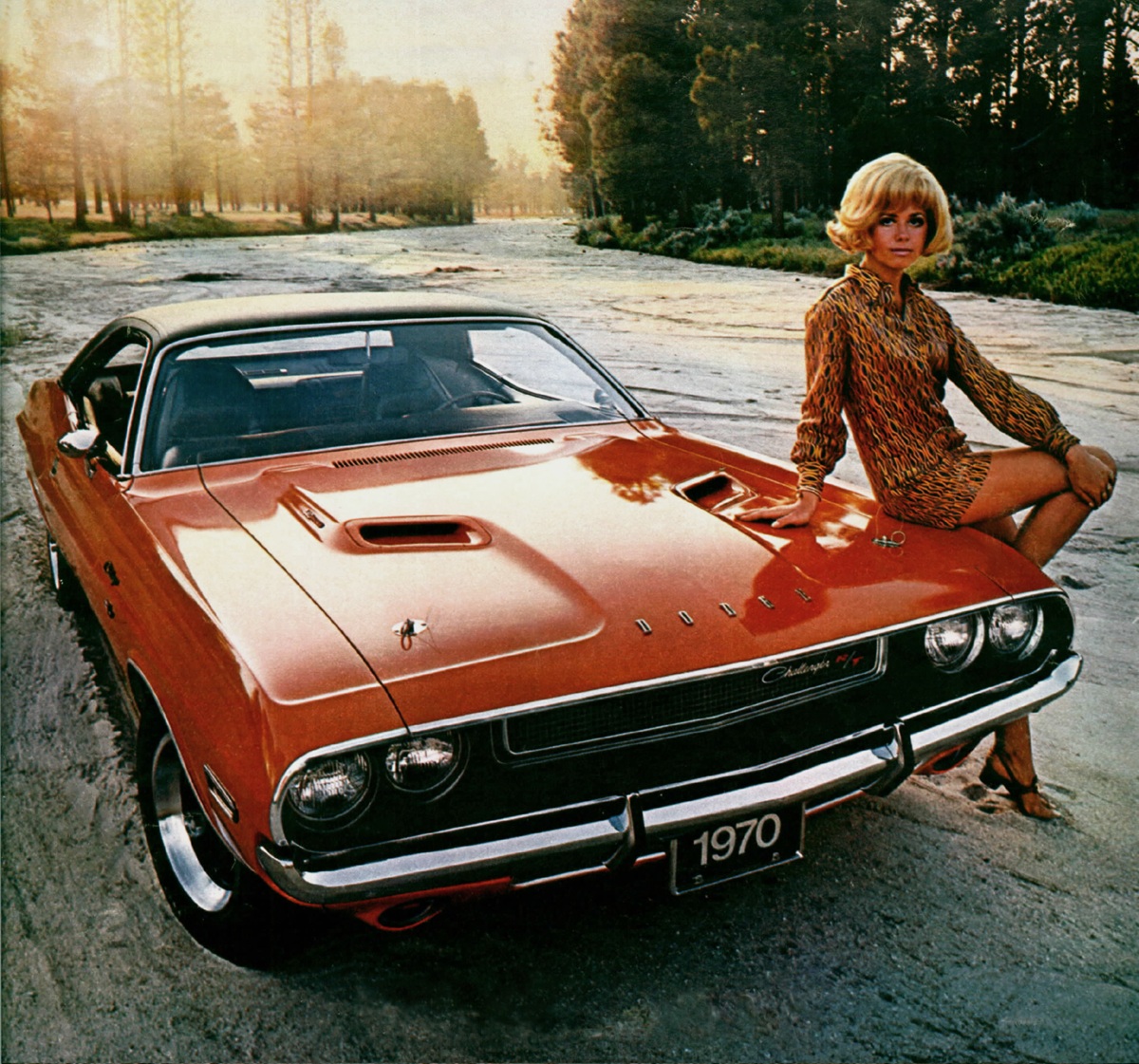 A woman in a patterned dress sits on the hood of an orange 1970 Dodge Challenger R/T parked on a sandy area with trees in the background at sunset.