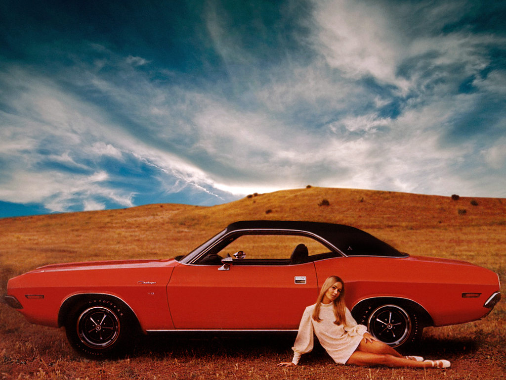 A young woman in a white dress sits on grass beside a bright orange classic muscle car in a hilly field under a dramatic blue sky with wispy clouds.