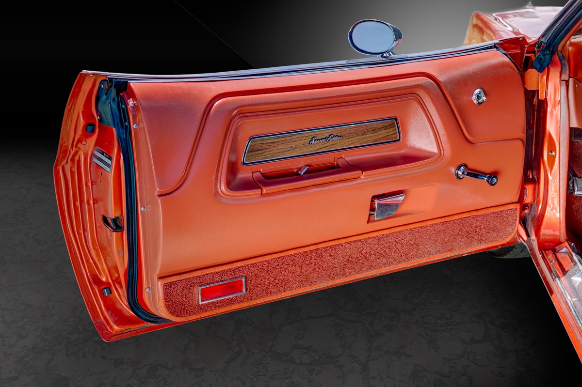 Close-up of an open vintage car door with orange interior, chrome handle, manual window crank, wood trim, and rectangular red reflector at the bottom. The background is dark and out of focus.