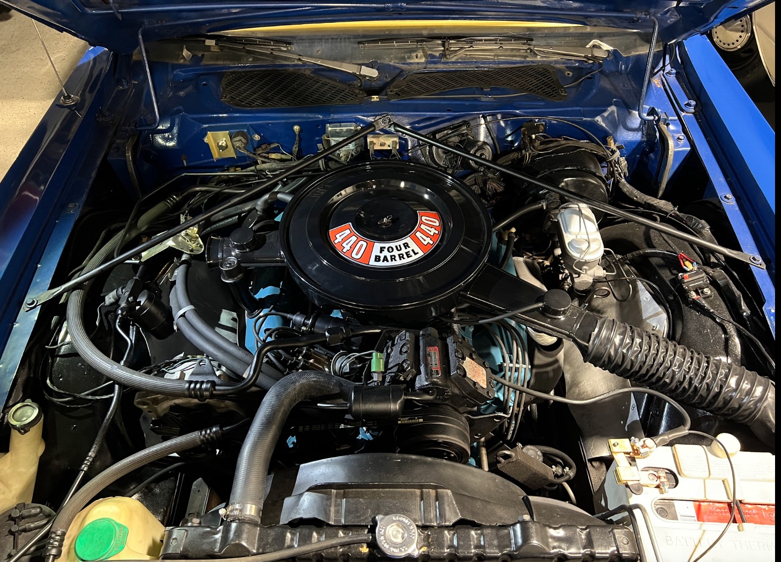 A close-up view of a classic car engine bay, featuring a prominently labeled 440 Four Barrel air cleaner, surrounded by various hoses, wires, and engine components inside a blue vehicle.