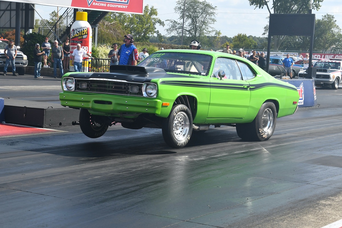 A bright green classic muscle car lifts its front wheels off the ground while launching down a drag strip, with spectators and other cars visible in the background.