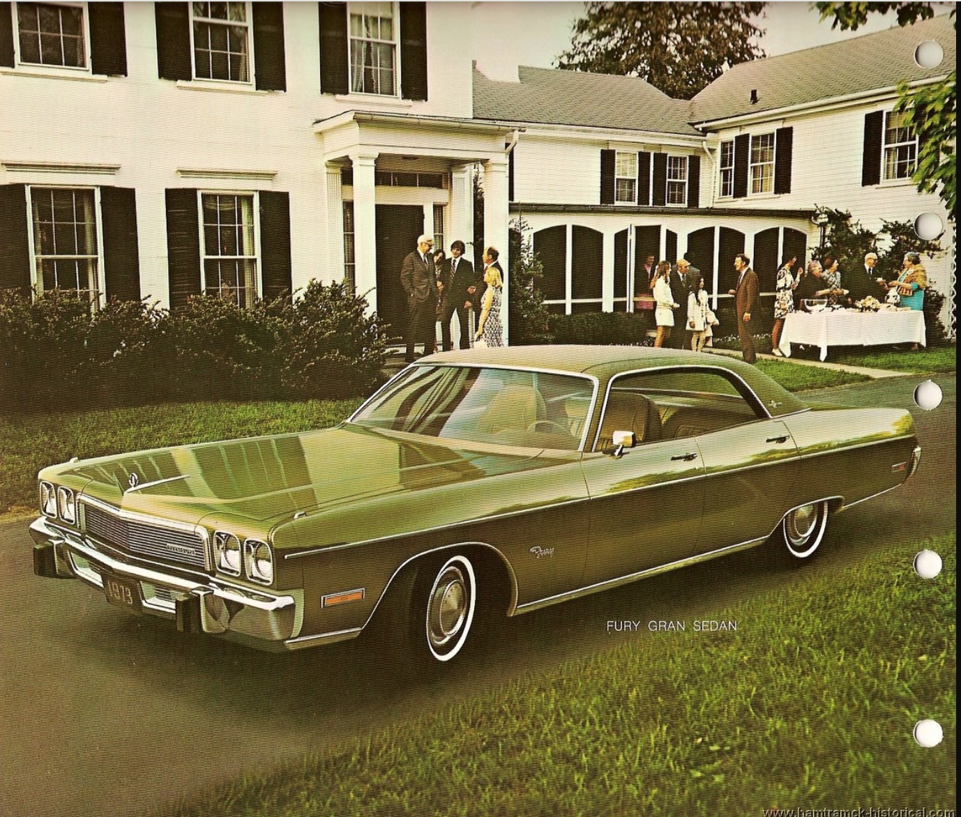 A vintage green 1973 Plymouth Fury Gran Sedan is parked on the lawn in front of a large white house, where people are dressed formally and socializing near a banquet table.
