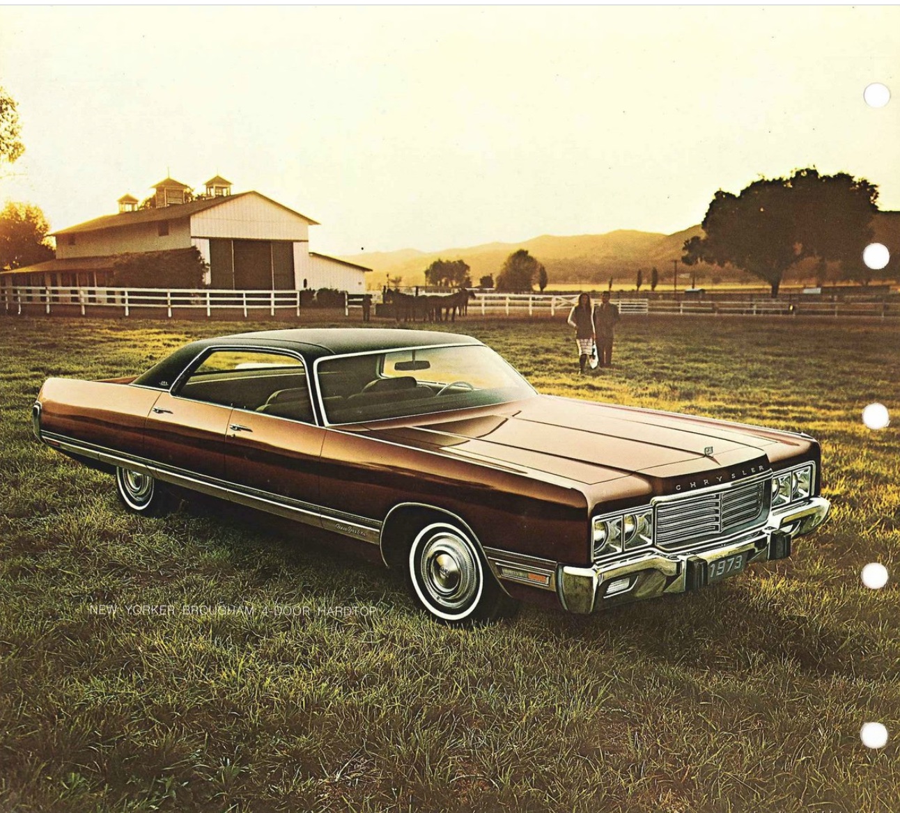 A vintage brown 1973 Chrysler New Yorker car parked on grass near a white fence, with a barn and trees in the background at sunset.