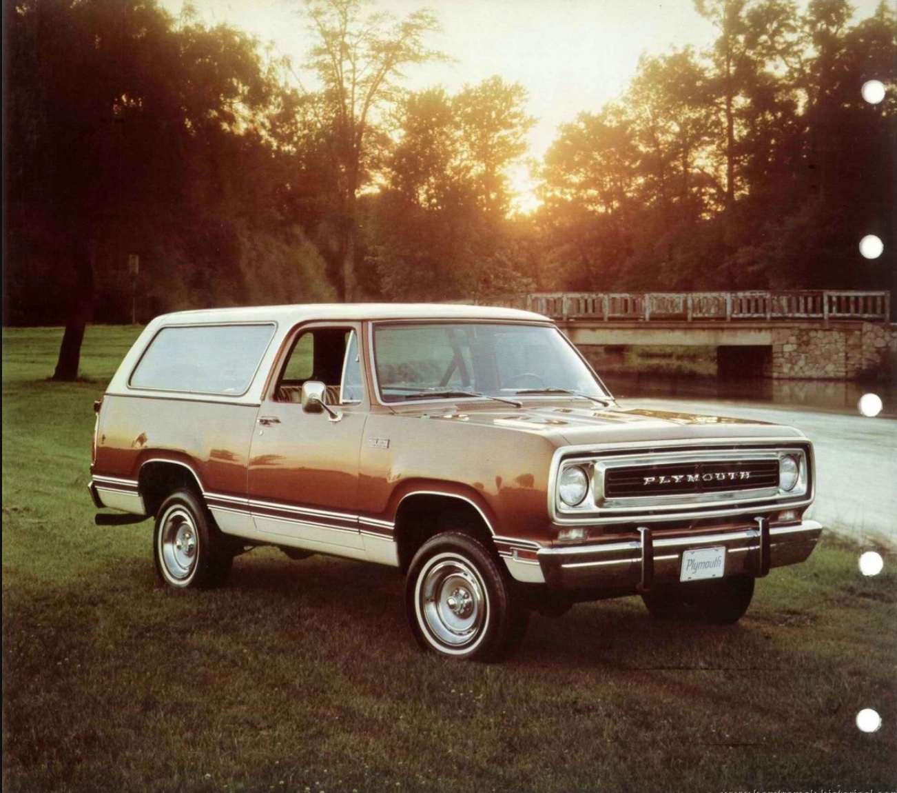 A vintage two-door Plymouth SUV with a brown and beige exterior is parked on grass near a river, with a stone bridge and trees in the background at sunset.