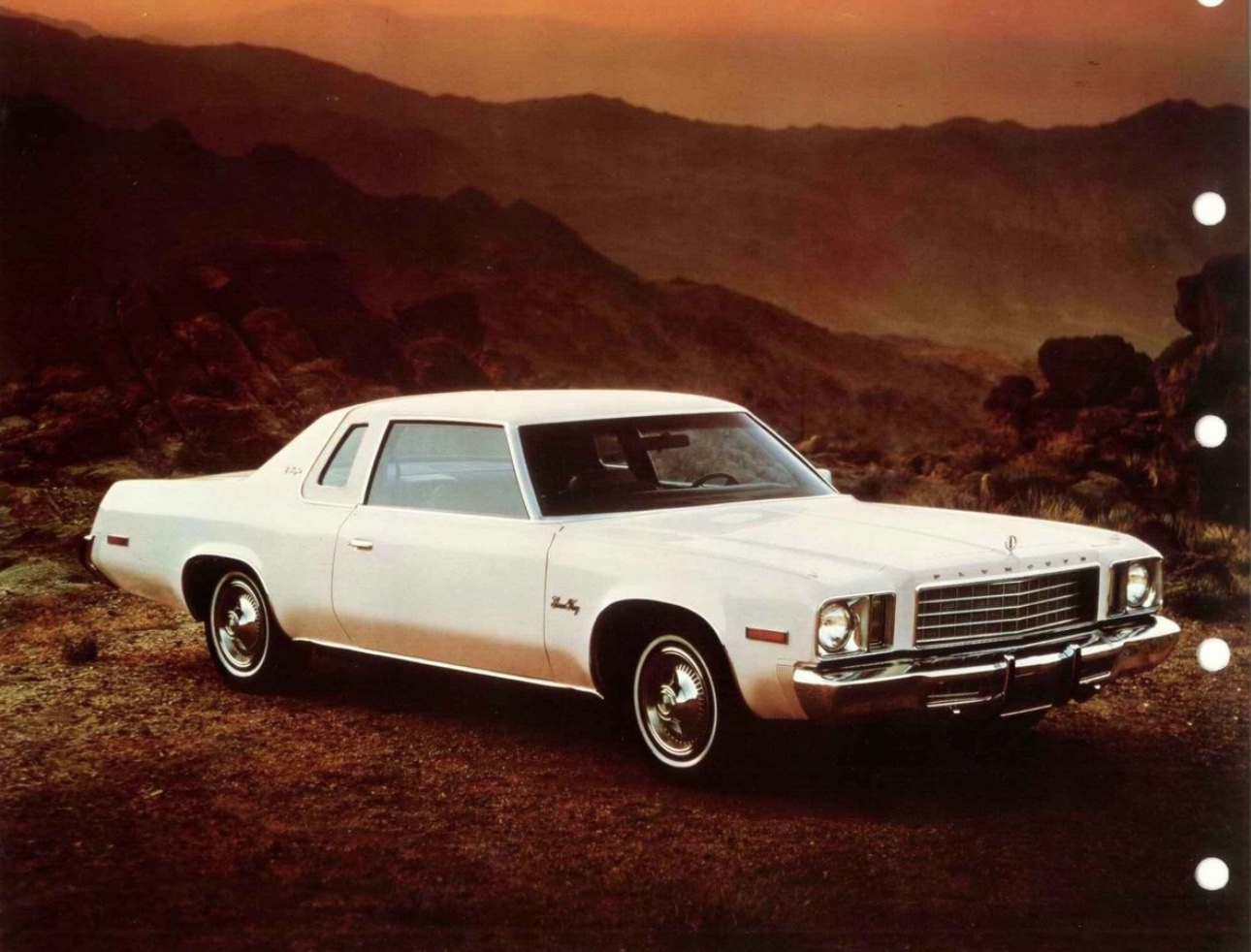 A white vintage two-door car is parked on a rocky landscape at sunset, with mountains and a dramatic sky in the background.