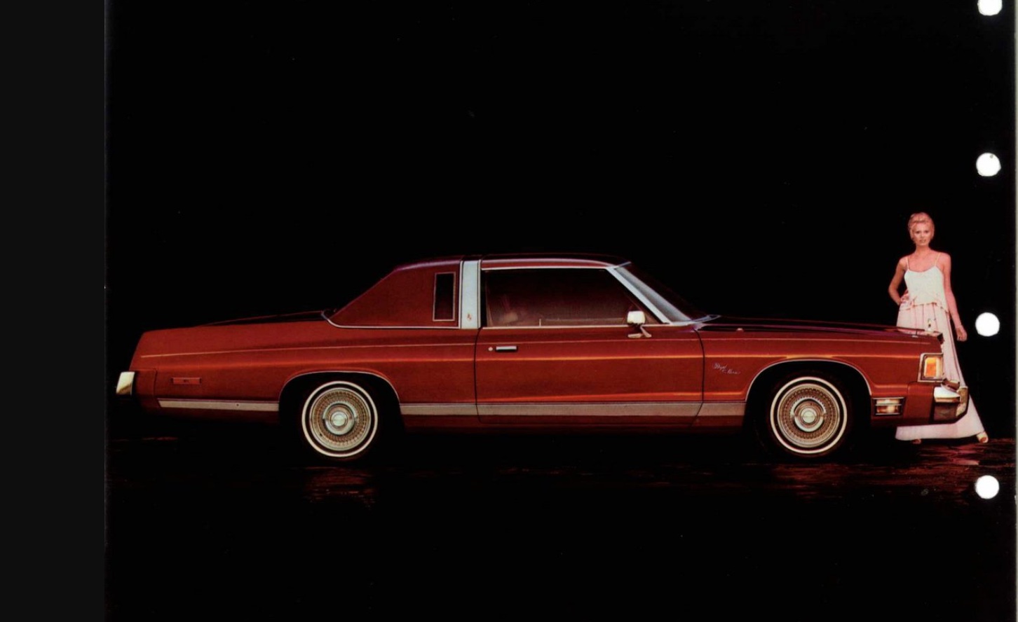A woman in a white dress stands next to a brown vintage two-door luxury car with a vinyl roof, set against a dark background.