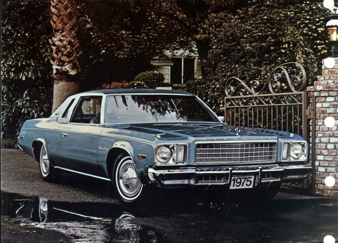 A vintage blue 1975 car is parked on a driveway in front of a wrought iron gate, with trees and a brick pillar in the background. The car has a boxy shape and chrome details.