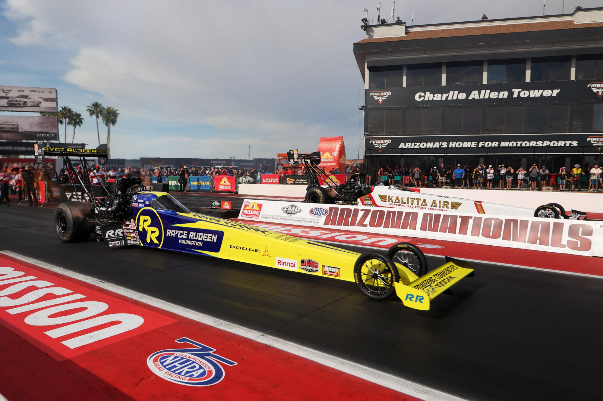 A yellow and blue dragster races down the NHRA Arizona Nationals track, with spectators and a large tower labeled Charlie Allen Tower in the background. Red banners and clear skies complete the lively scene.