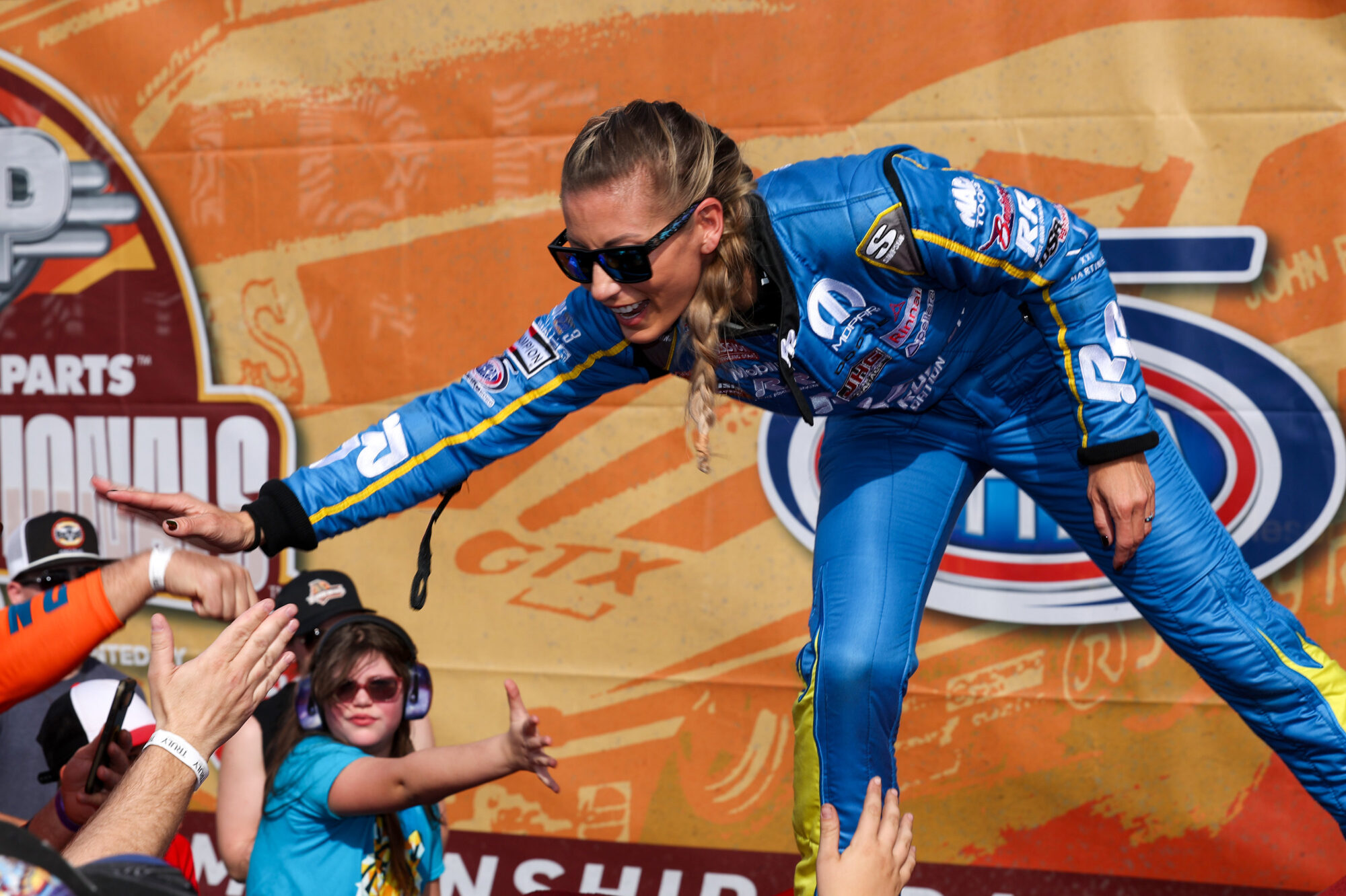 A smiling race car driver in a blue suit and sunglasses leans forward to high-five enthusiastic fans, including a child wearing ear protection, against a colorful event backdrop.