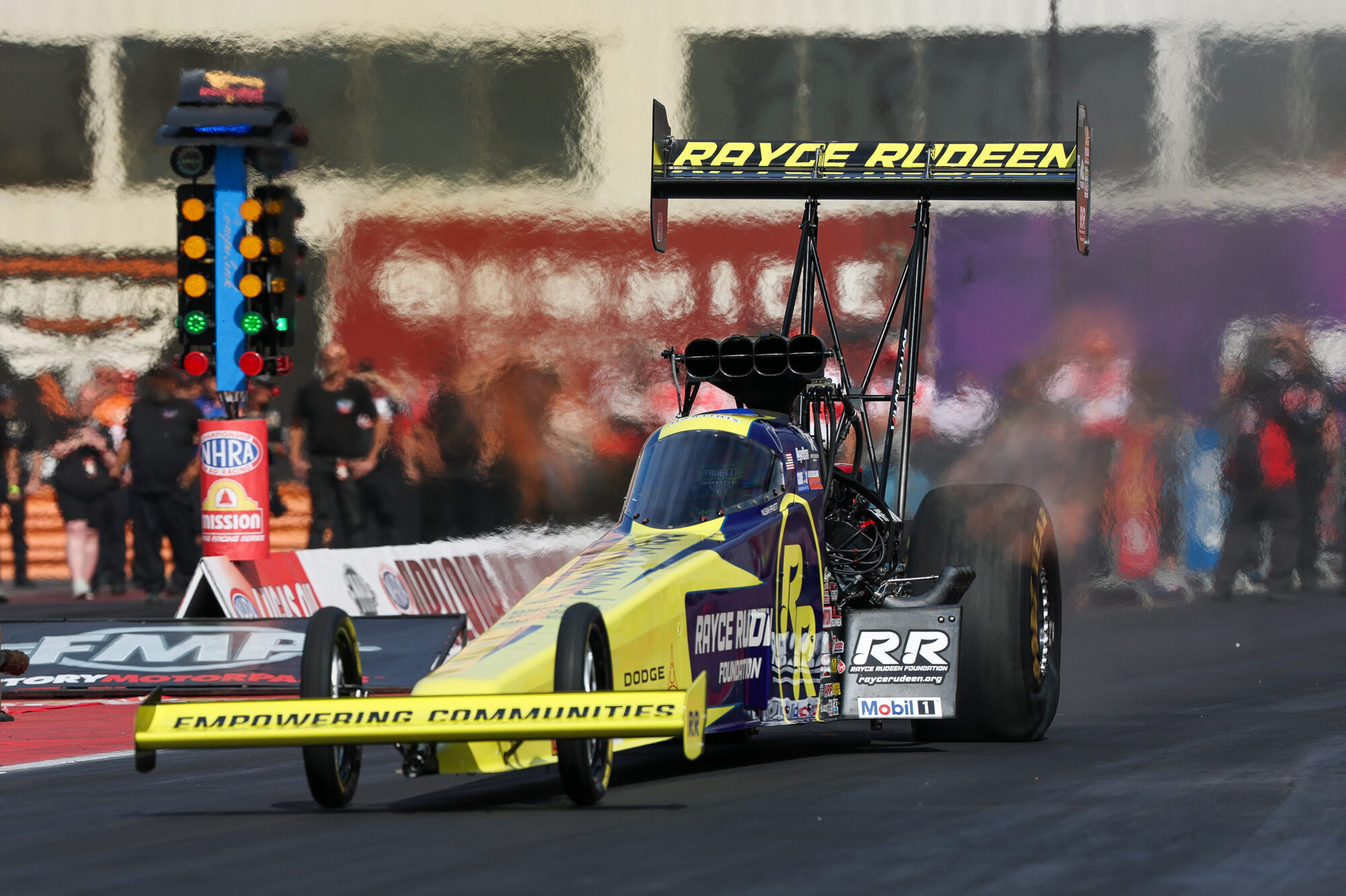 A yellow and purple drag racing car accelerates down a racetrack, producing smoke from its rear tires. The rear wing displays “RAYCE RYDEEN.” Team members and officials stand near the track in the background.