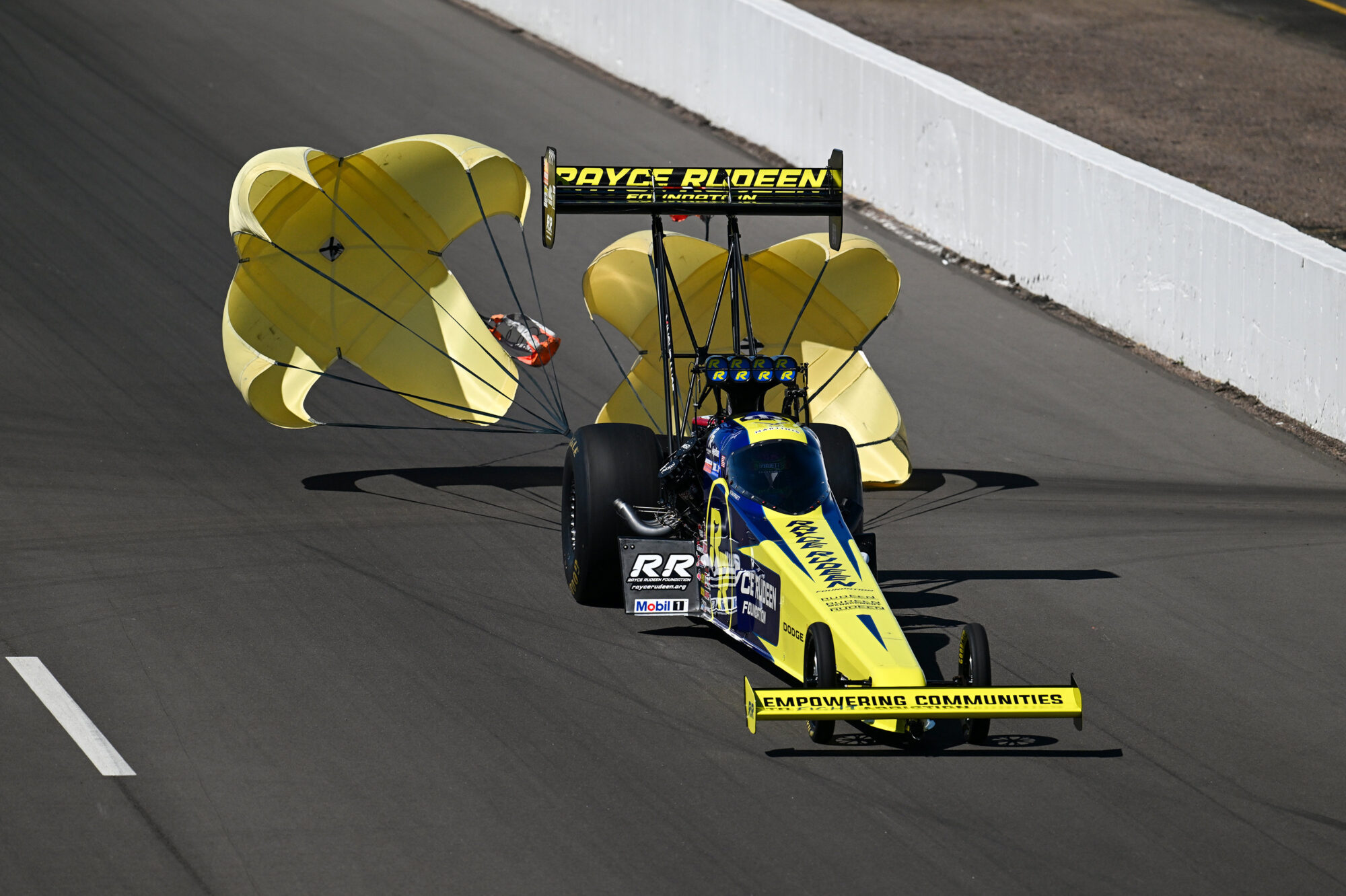 A yellow and black dragster deploys two large yellow parachutes as it slows down on a racetrack, with a white barrier and asphalt visible in the background.