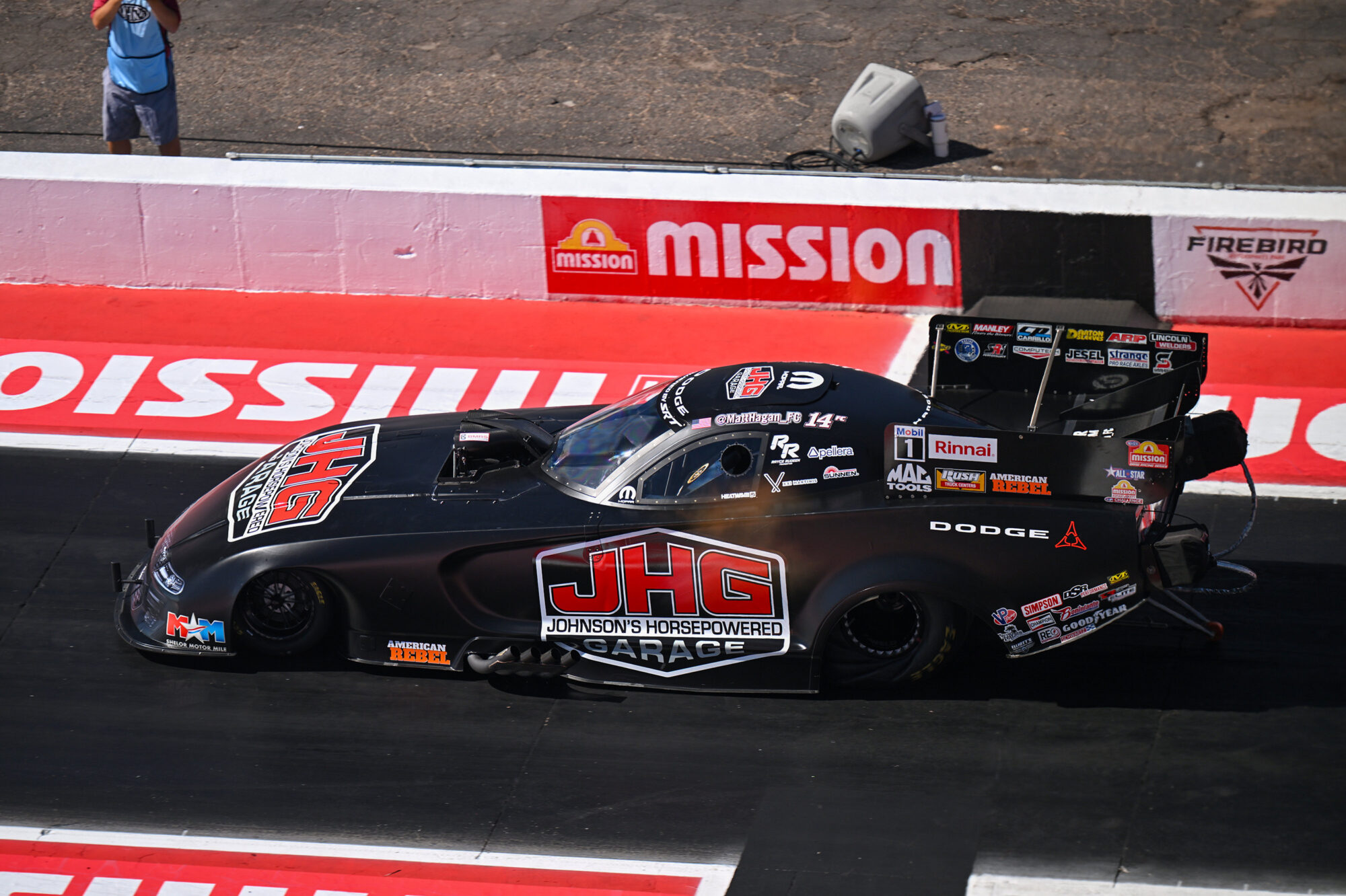 A black Dodge drag racing car, covered in sponsor logos including a large JHG on the hood, speeds down a racetrack at Firebird Raceway, with Mission-branded barriers in the background.