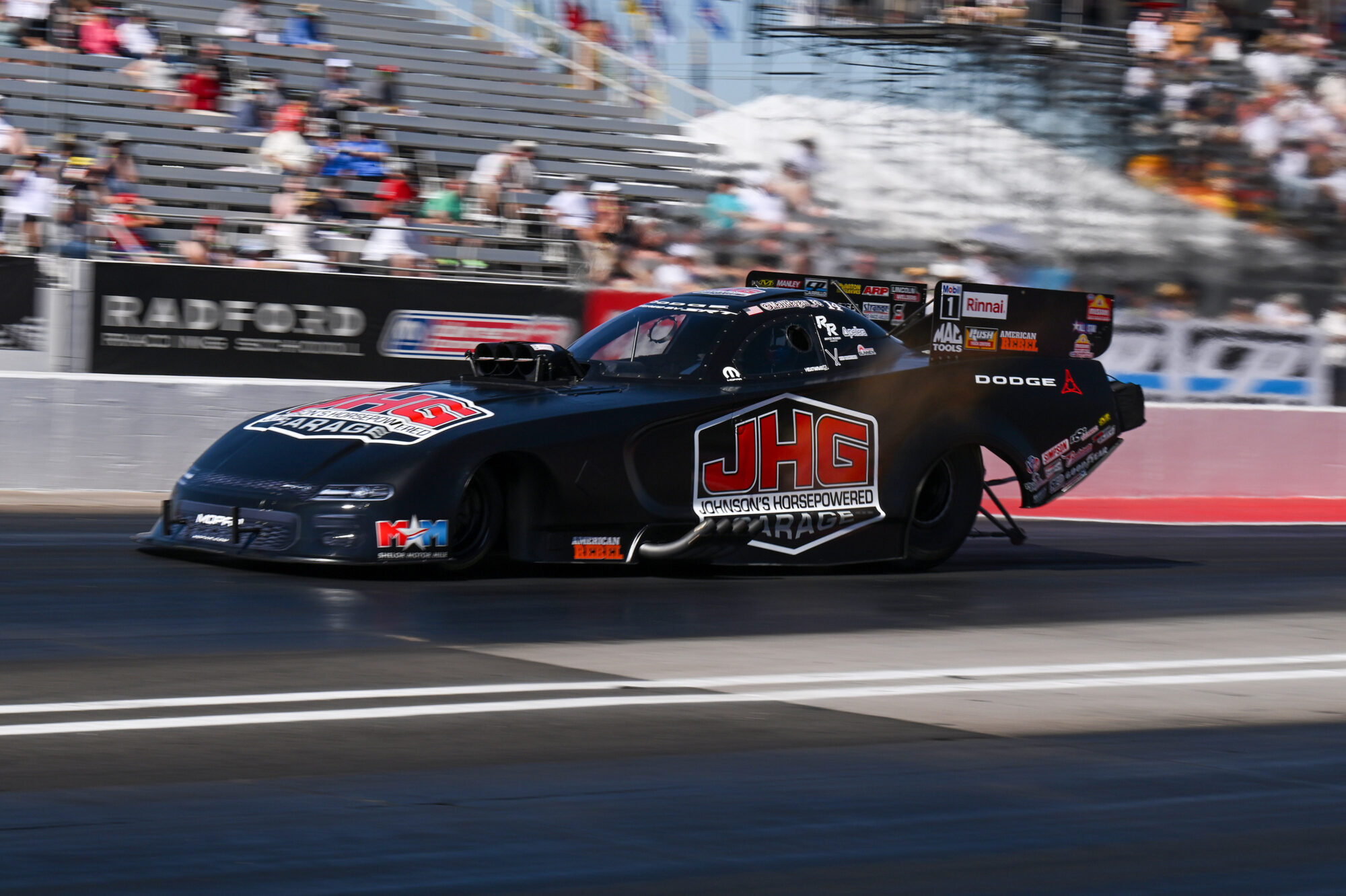 A black drag racing car with JHG branding speeds down a racetrack, leaving blurred motion behind. Spectators fill the stands in the background under bright daylight.