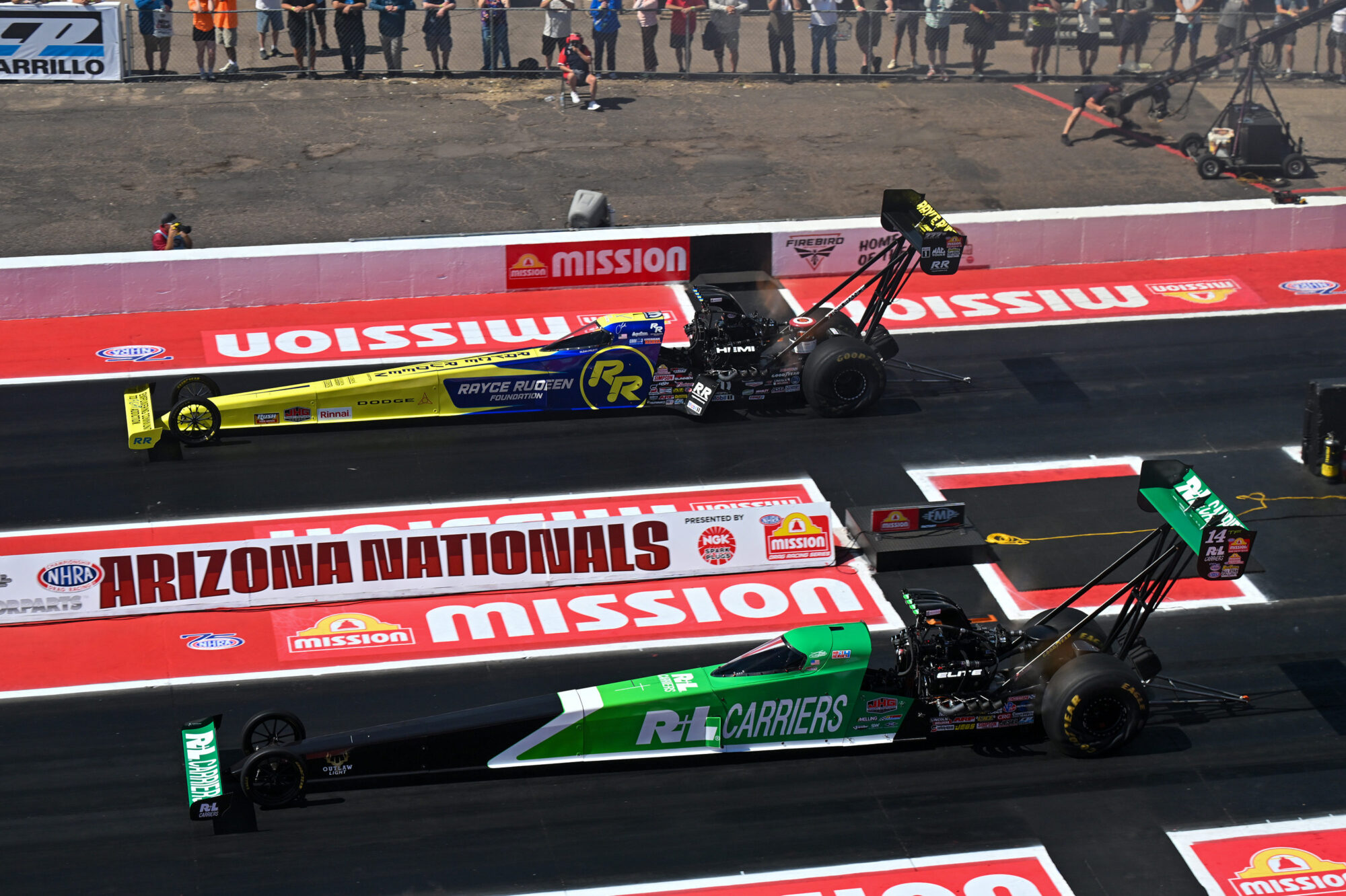 Two drag racing cars, one black and blue, the other green and black, line up side by side at the start of a race on a red and white track at the Arizona Nationals event, with spectators watching in the background.
