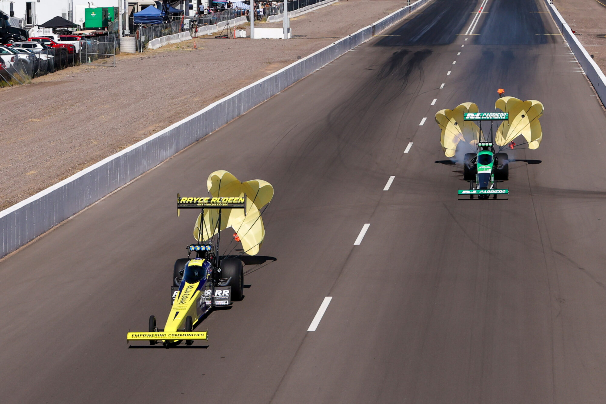 Two drag racing cars slow down on a racetrack with yellow parachutes deployed behind them; a fenced area and tents are visible to the left, and tire marks are on the road.