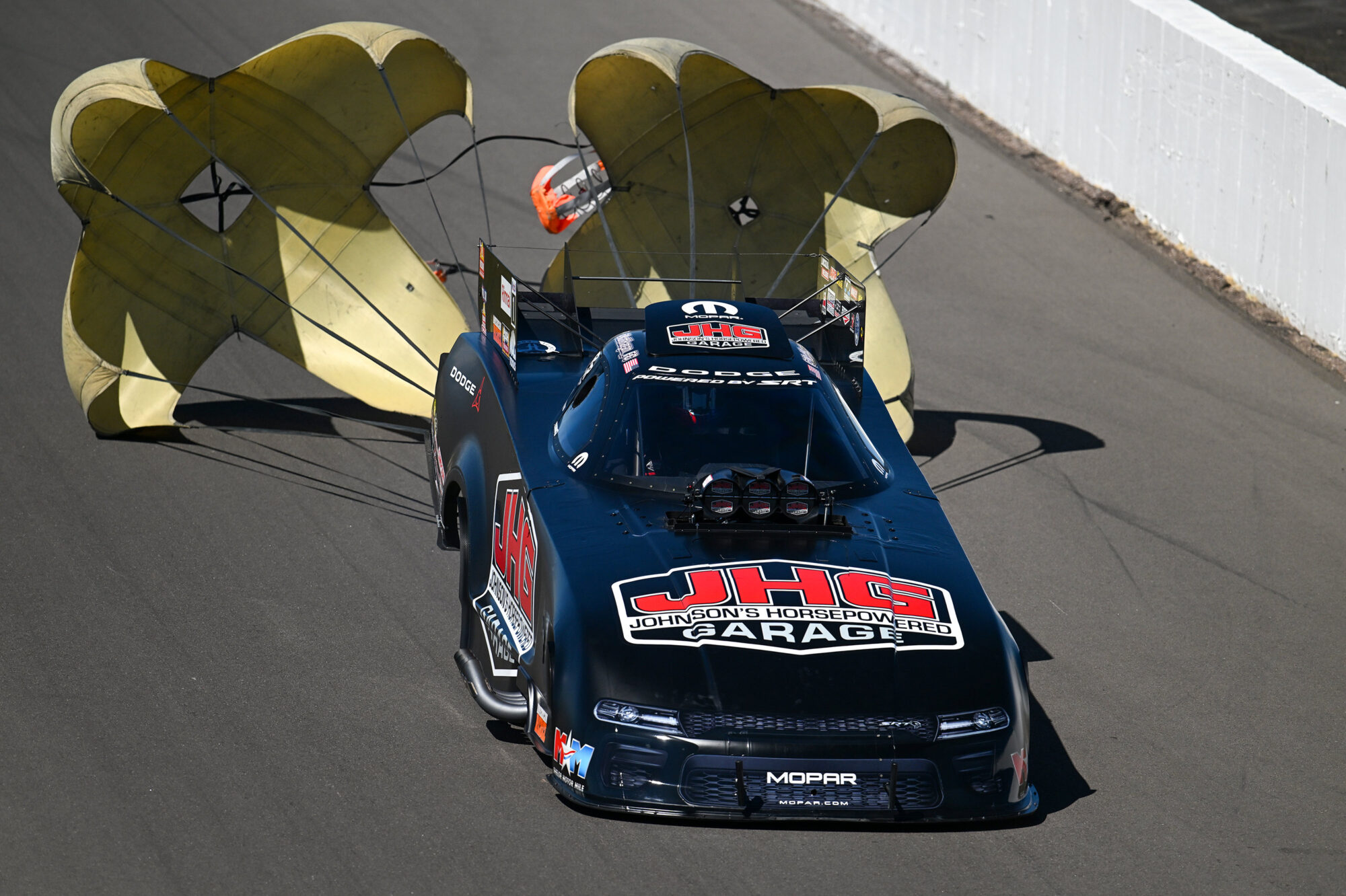 A black drag racing car with “JHG Johnson’s Horsepowered Garage” on the hood deploys two large yellow parachutes as it slows down on a racetrack.
