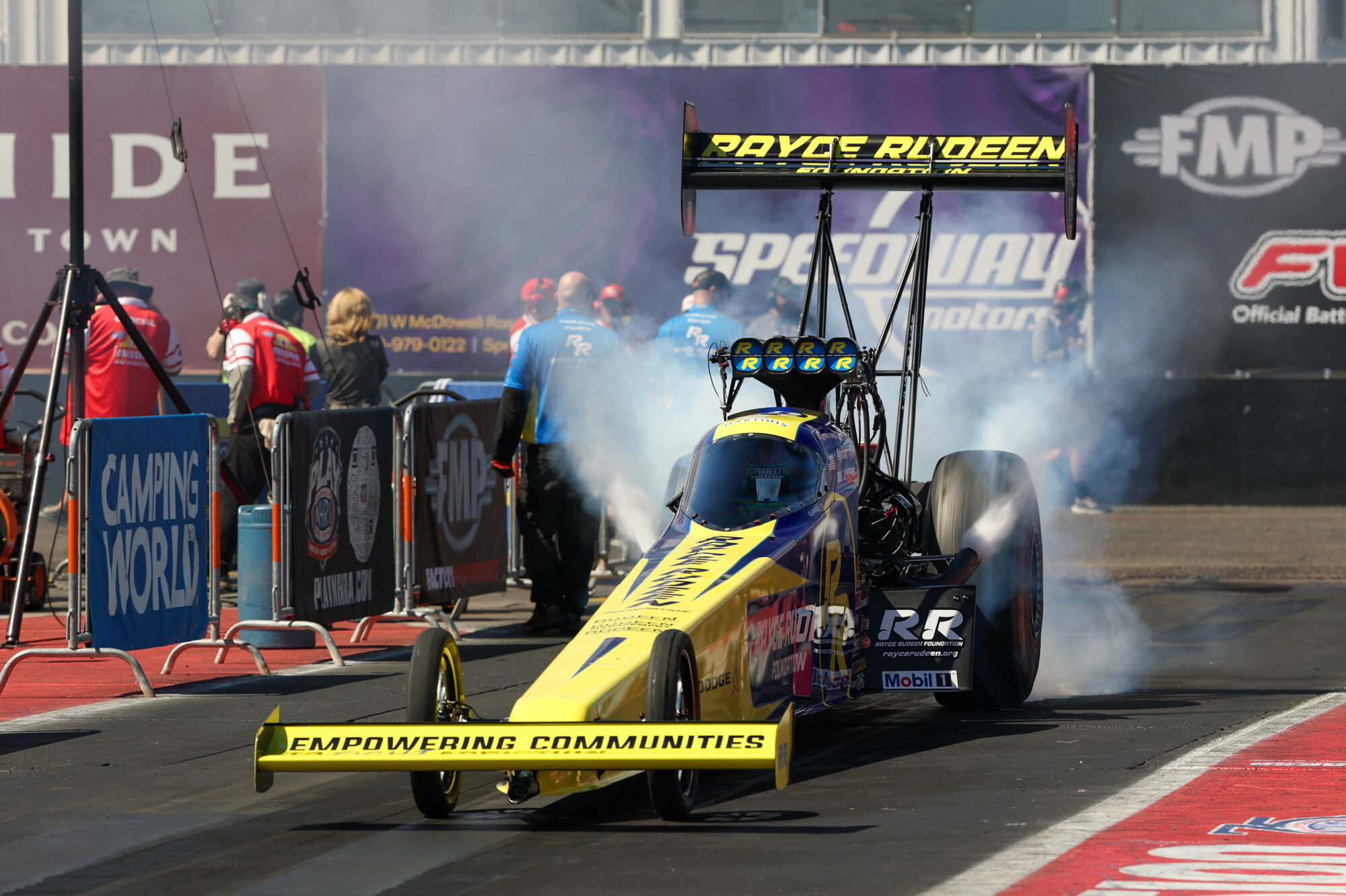A yellow and black dragster with “Empowering Communities” on its front wing releases smoke as it prepares to race on a drag strip. Crew members and spectators stand by, with banners and sponsor logos visible in the background.