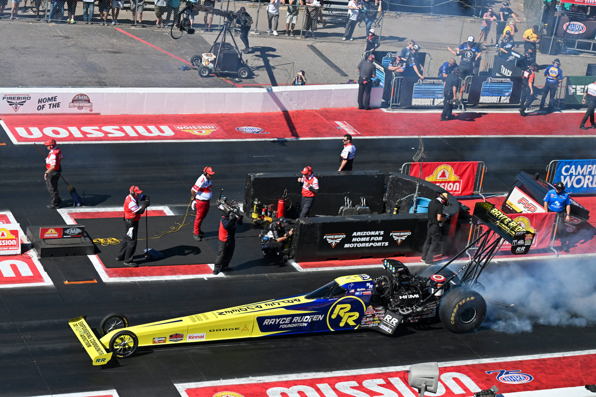 A drag racing car with a yellow and blue livery performs a burnout at a racetrack, surrounded by crew members in red uniforms and smoke, while spectators watch from above.