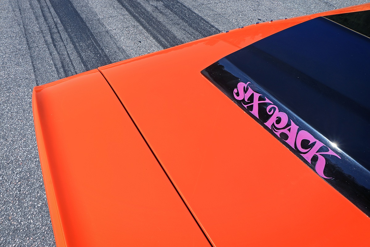 Close-up of the orange hood of a car with a black section that has SIX PACK written in pink letters. Faint tire marks are visible on the gray asphalt in the background.