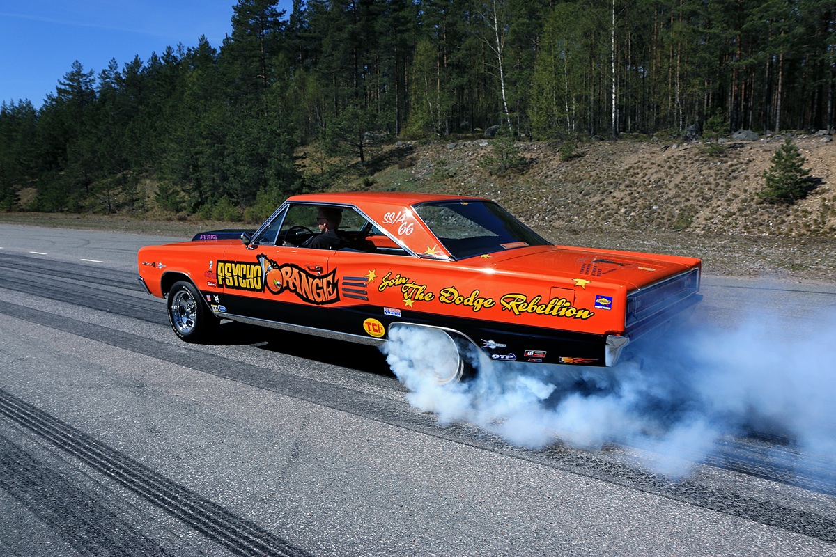 A bright orange classic muscle car performs a burnout on an open road, producing thick white smoke from its rear tires, with trees and a hillside in the background.