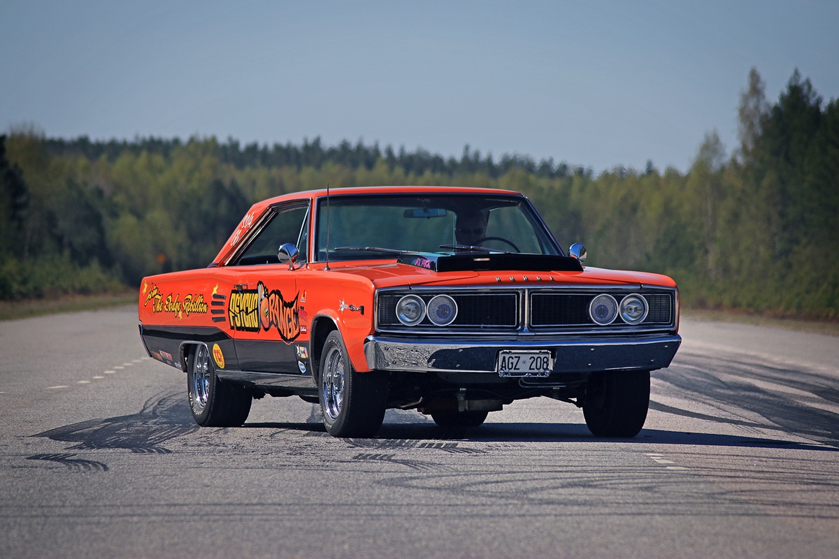 A bright orange vintage muscle car with graphics on the side is performing a burnout on an empty road, leaving tire marks. Green trees and blue sky are visible in the background.