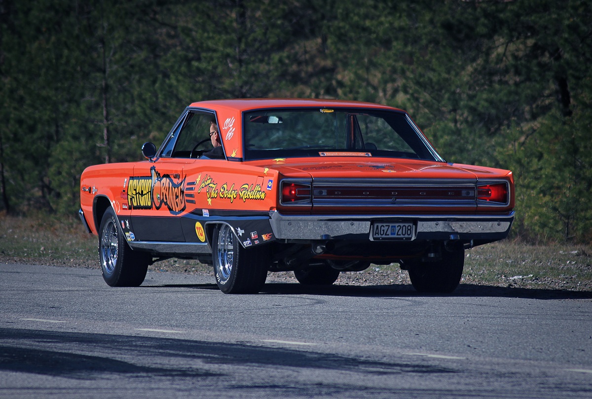 A classic orange muscle car with custom decals and shiny chrome wheels is parked on a road, with trees visible in the background. The cars rear and side are in view, showing its vintage design and bold graphics.