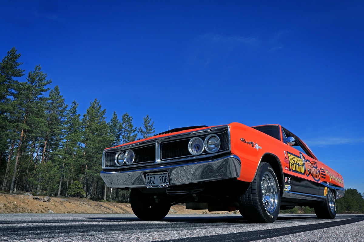 Low-angle view of a bright orange and black vintage muscle car parked on a road, with trees and a clear blue sky in the background. The car has shiny chrome wheels and Show Bus painted on the side.