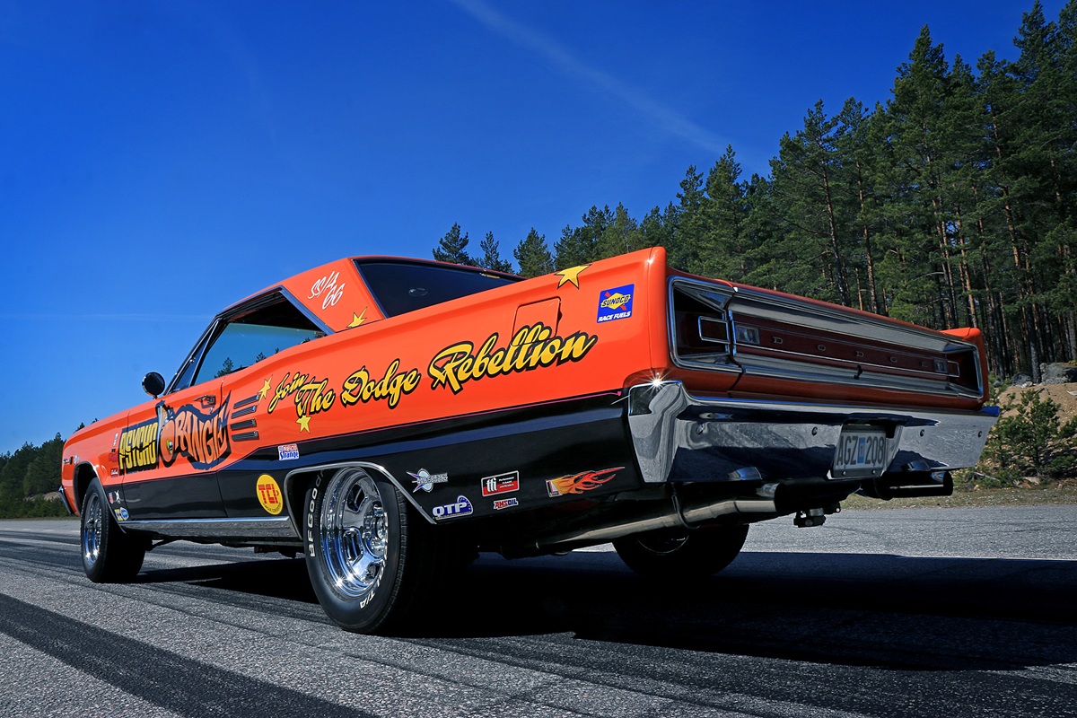 A bright orange vintage muscle car with racing decals and “The Dodge Rebellion” written on the side is parked on an asphalt road, surrounded by trees and a clear blue sky.