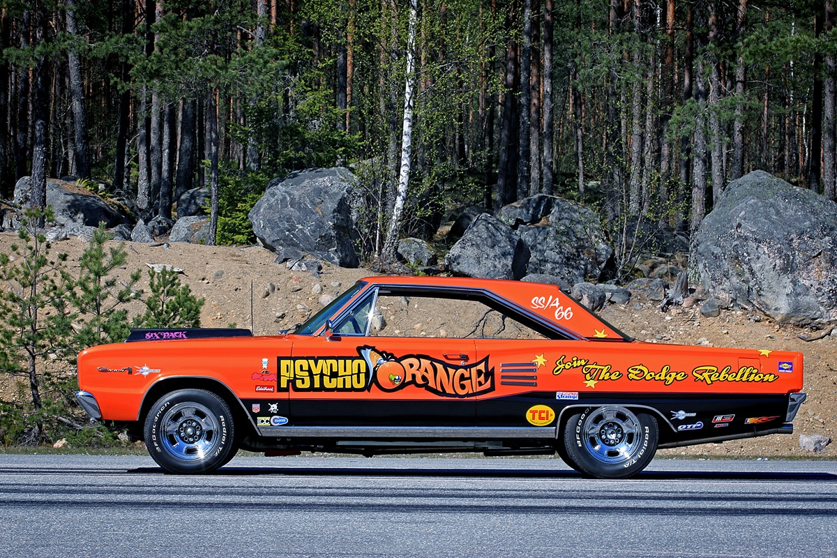 A vintage orange drag racing car with Psycho Orange and Join the Dodge Rebellion painted on the side, parked on a road with rocks and pine trees in the background.
