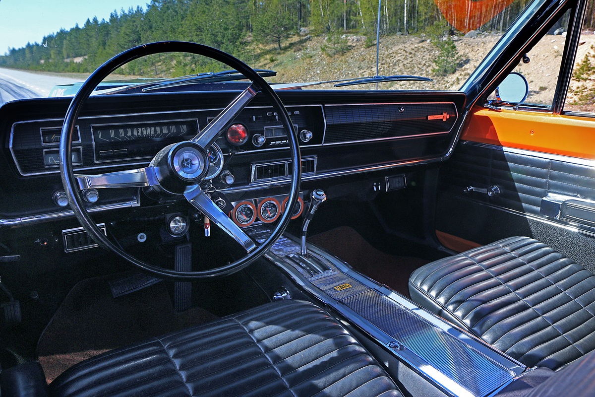 Classic car interior featuring a large black steering wheel, vintage dashboard with analog gauges, black leather seats, and silver console accents. Sunlight highlights the orange trim and outside trees are visible through the window.