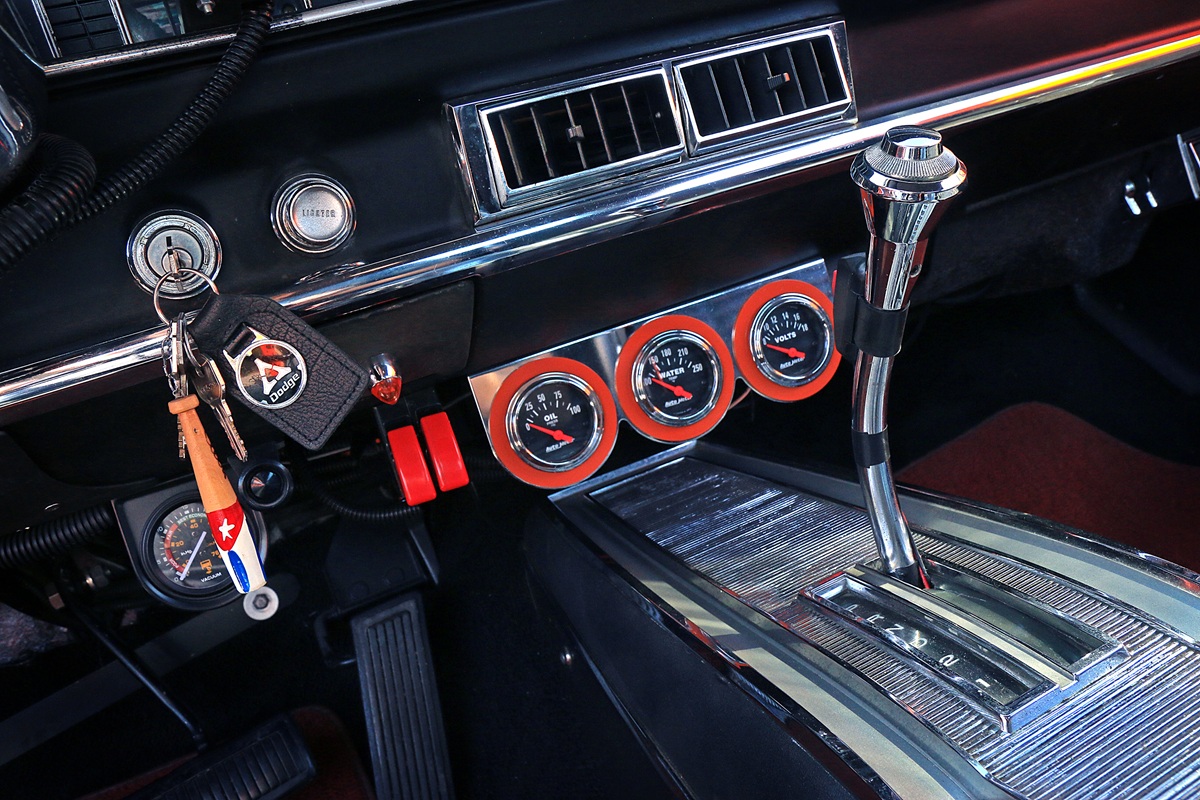 Close-up of a classic car interior showing a gear shift, dashboard gauges, air vents, and keys hanging with a Cuban flag keychain. Chrome and red accents are visible.