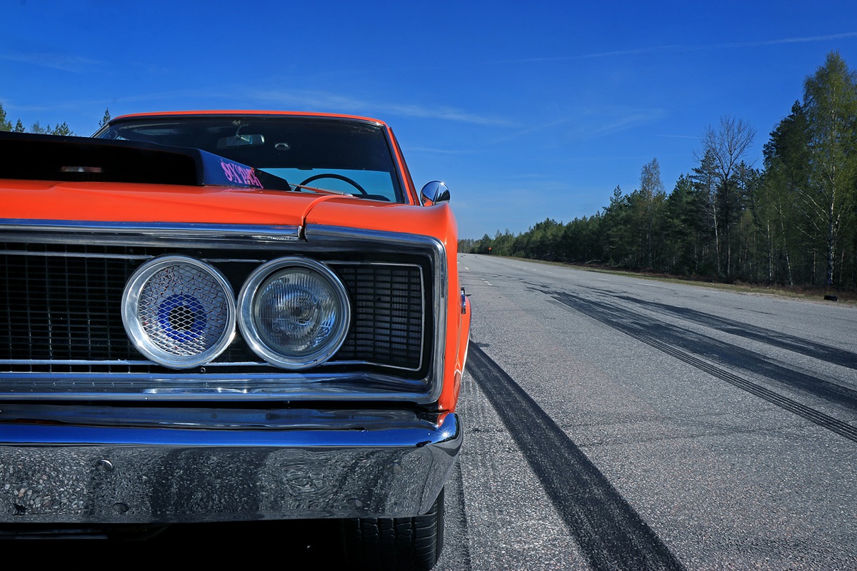 Close-up of the front left side of a classic orange car parked on an empty road with visible tire marks, surrounded by trees and under a clear blue sky.