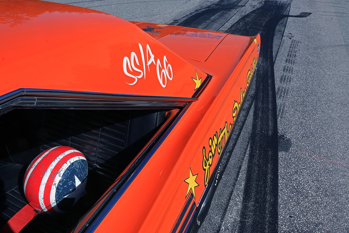 Close-up of an orange vintage race car with yellow stars and text, and a helmet with an American flag design on the drivers seat. The car is on asphalt with visible tire skid marks.