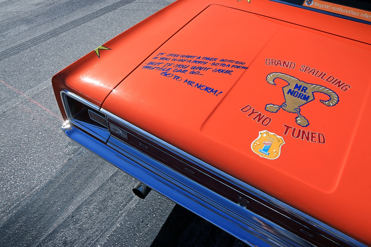 Bright orange muscle car trunk with blue MR. NORM logo, Grand Spaulding Dyno Tuned text, and a humorous quote; chrome bumper and license plate visible under clear daylight.