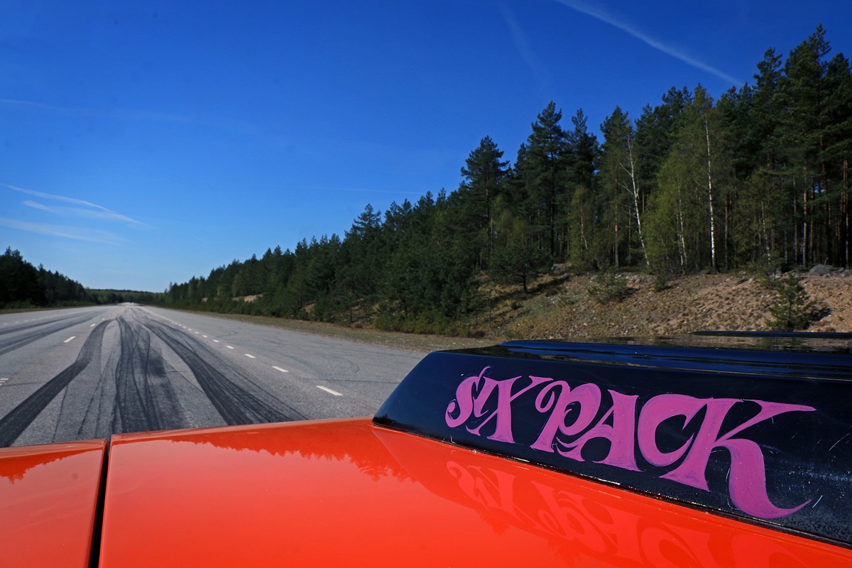 A bright orange car with a Six Pack decal on its hood is parked on an empty road bordered by pine trees and under a clear blue sky. Tire marks are visible on the road.