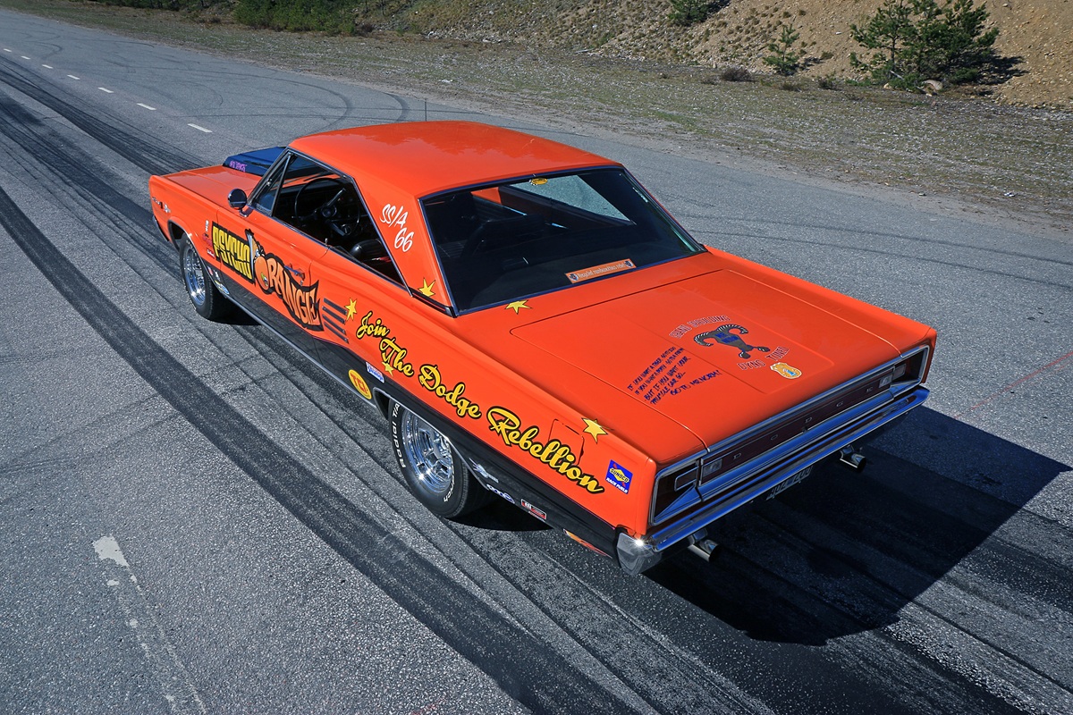 A bright orange vintage muscle car with “HEMI ROAD RUNNER” and “Join the Dodge Rebellion” decals is parked on a road with visible tire marks, viewed from above and behind. The car has chrome wheels and a blue interior.